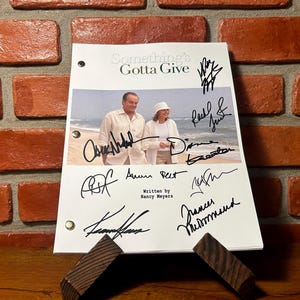 Puede incluir: Un guion de la película "Something's Gotta Give" con firmas. El guion está sostenido por un soporte de madera. El título y una foto de los actores son visibles. El fondo es una pared de ladrillos.