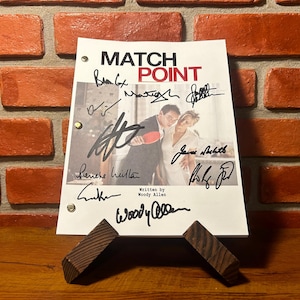 May include: A signed script for the film "Match Point" by Woody Allen. The script features the film's title in red and black, a movie still, and multiple signatures. It is displayed on a wooden stand against a brick wall.
