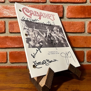 Puede incluir: Un guion del musical "Cabaret" con múltiples firmas, exhibido en un soporte de madera. El título "Cabaret" está en escritura roja. El guion presenta una foto en blanco y negro de artistas.