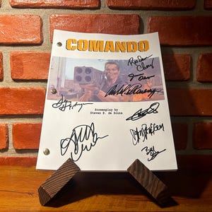 May include: A script for the movie "Commando" with the title at the top in yellow letters. The script is signed by several people. The script is propped up on a wooden stand against a brick wall.