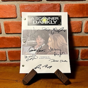 Può includere: Una sceneggiatura firmata del film "A Scanner Darkly" con Keanu Reeves e Winona Ryder. Il titolo è in alto, con firme sulla pagina. La sceneggiatura è tenuta da un supporto in legno contro un muro di mattoni.