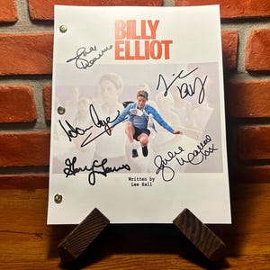 May include: A script for "Billy Elliot" with the title in red and the image of a boy jumping. The script is signed by multiple people. The script is held up by a wooden stand, with a brick wall in the background.