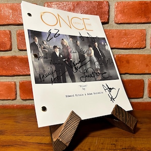 Peut inclure: Un script signé de la série télévisée "Once Upon a Time", présenté sur un présentoir en bois. Le script porte le logo de l'émission et le titre "Pilot" par Edward Kitsis et Adam Horowitz. Le fond est un mur de briques rouges.