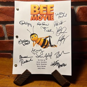 Könnte beinhalten: Ein Drehbuch für den Animationsfilm "Bee Movie" mit dem Titel in Orange und Gelb. Das Drehbuch zeigt eine Zeichentrickbiene und mehrere Unterschriften. Das Drehbuch wird von zwei Holzstützen gehalten.
