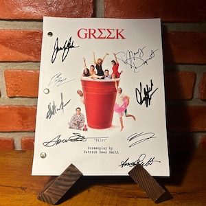May include: A signed "Greek" screenplay titled "Pilot" by Patrick Sean Smith. The cover features a red cup with people inside, surrounded by signatures. The script is displayed on a wooden stand against a brick wall.