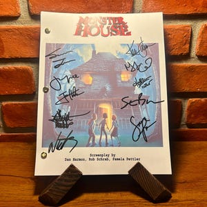Puede incluir: Un guion firmado de la película de animación "Monster House" con el título de la película en rojo y azul. El guion está sujeto por un soporte de madera y presenta firmas y los nombres de los guionistas.