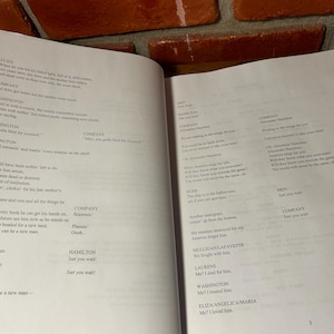 May include: Open book with printed text on white pages. The text appears to be a script or play, with character names and dialogue. The pages are slightly yellowed, suggesting age. The book is open on a dark surface.