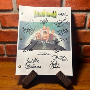 Puede incluir: Una hoja de papel blanca con el título de la película "ParaNorman" e ilustraciones de personajes, con múltiples firmas en tinta negra. El guion está apoyado en un soporte de madera contra una pared de ladrillo.