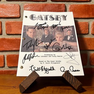 Könnte beinhalten: Ein signiertes Drehbuch von "Gatsby" liegt auf einem Holzständer. Der Titel steht oben, darunter die Unterschriften der Darsteller. Das Bild zeigt ein Foto der Besetzung und den Text "Drehbuch von Baz Luhrmann und Craig Pearce."