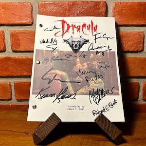 May include: A film script for "Dracula" featuring numerous signatures, presented on a wooden stand. The title is in red, with a bat emblem. The backdrop is a red brick wall.