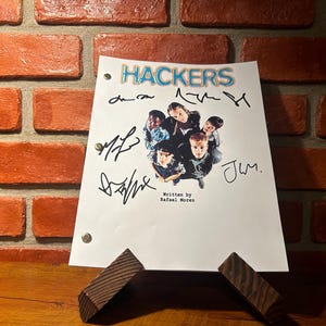 Può includere: Una sceneggiatura bianca con il titolo "Hackers" e firme, sostenuta da un supporto in legno. La sceneggiatura presenta una foto di gruppo del cast. Lo sfondo è un muro di mattoni.