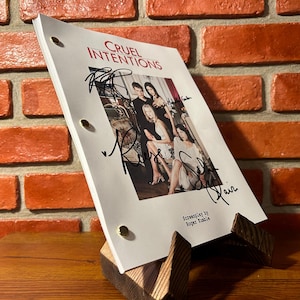 May include: A script for the movie "Cruel Intentions" is displayed on a wooden stand. The script has the movie title and a photo of the cast. The script is signed by the cast. The background is a brick wall.