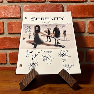 May include: A film script for "Serenity" featuring signatures. The title "Serenity" is at the top, with a cast photo. The script is supported by two small wooden props, set against a brick wall.