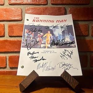May include: A script for "The Running Man" movie, featuring signatures. The title is in red, with a movie scene image. The script is held by a wooden stand against a brick wall.
