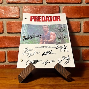 May include: A white sheet with the title "PREDATOR" in red letters, featuring an image of a man holding a gun. The sheet is signed by multiple people and is propped up by two wooden supports.