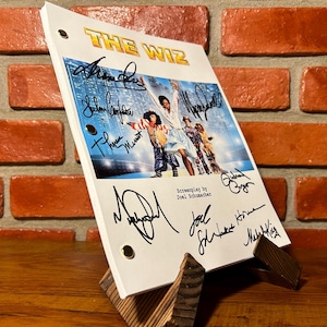 Op de afbeelding: Een wit script voor "The Wiz" met meerdere handtekeningen, weergegeven op een houten standaard. Het script bevat een kleurenfoto van de cast van de film. De titel "The Wiz" is in gouden letters.