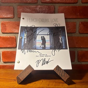 May include: A white script with the title "PUNCH-DRUNK LOVE" and signatures, displayed on a wooden stand. The script features a still from the film and is written by Paul Thomas Anderson. The background is a brick wall.