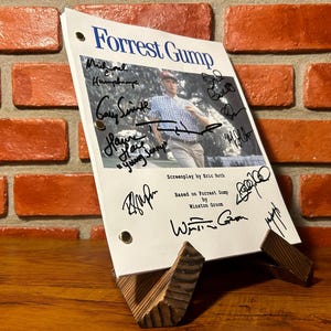 May include: A script for the movie "Forrest Gump" with the title in blue and signatures. The script is propped up on a wooden stand against a brick wall. The script includes the text "Screenplay by Eric Roth" and "Based on Forrest Gump by Winston Groom."