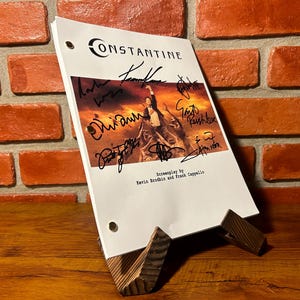 May include: A script for the movie "Constantine" with multiple signatures, displayed on a wooden stand. The script cover features the movie title and a promotional image. The screenplay is by Kevin Brodbin and Frank Cappello.