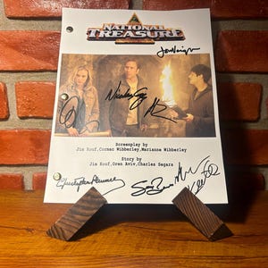 May include: A signed script from the movie "National Treasure" featuring the film's title and cast. The script is displayed on a wooden stand against a brick wall background.