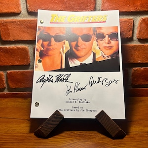 May include: A script for "The Grifters" movie, featuring a movie poster image and signatures. The title is in yellow and red. The script is held up by two wooden supports, set against a brick wall and wooden surface.
