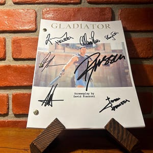 May include: A white script from the movie "Gladiator" with multiple signatures, including "Russell". The script is propped up on a wooden stand against a red brick wall.