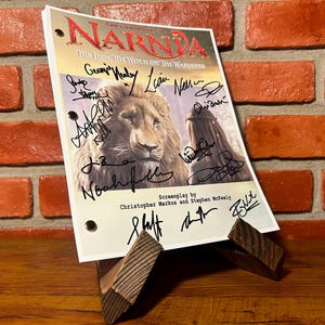 May include: A script for "The Lion, the Witch, and the Wardrobe" is displayed on a wooden stand. The script features the movie title and images of a lion and a woman. The script is signed by multiple people.