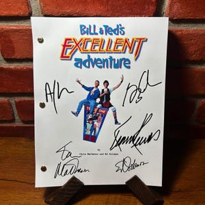 May include: A white paper featuring the title "Bill & Ted's Excellent Adventure" in blue, red, and yellow. The paper has illustrations of two men and is signed by several people. The paper is held up by a wooden stand.
