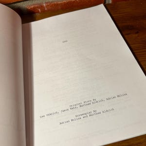 May include: Open script pages with text: "Original Story By" followed by names, and "Screenplay By" followed by names. The pages are white, with black text, and are on a wooden surface.
