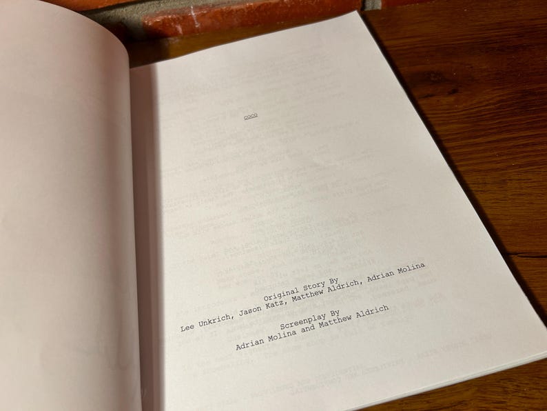 May include: Open script pages with text: "Original Story By" followed by names, and "Screenplay By" followed by names. The pages are white, with black text, and are on a wooden surface.