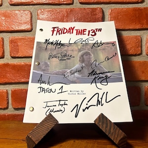 May include: A signed script page from "Friday the 13th" featuring the film's title in red and multiple signatures. The script page is displayed on a wooden stand against a brick wall.