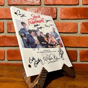 May include: A signed script for "Shock Treatment", displayed on a wooden stand. The script includes a cast photo and signatures. The background is a brick wall and a wooden surface.