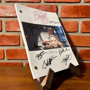 May include: A script for the movie "Brazil" with multiple signatures, displayed on a wooden stand. The title "Brazil" is written in pink script. The script is held together with brass fasteners. The background is a brick wall.