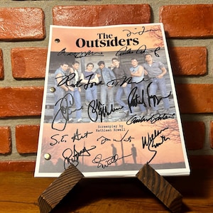 May include: A printed page from "The Outsiders" screenplay, featuring a group photo and signatures. The page is propped up on a wooden stand against a brick wall. The title "The Outsiders" is at the top.