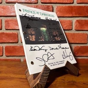 Könnte beinhalten: Ein Filmskript für "Prince of Darkness" mit Unterschriften, präsentiert auf einem Holzständer. Der Titel steht oben, darunter die Namen der Schauspieler und der Crew. Das Skript steht auf einem Holzständer vor einer Backsteinmauer.
