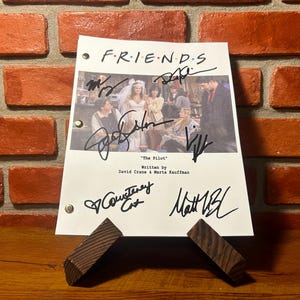 May include: A signed script from the TV show "Friends", titled "The Pilot". The script is supported by a wooden stand, set against a brick wall. The title "FRIENDS" is visible at the top of the page.
