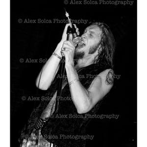 May include: Black and white photograph of a person singing into a microphone. The person is wearing a vest and has long hair. The image includes the text "© Alex Solca Photography".