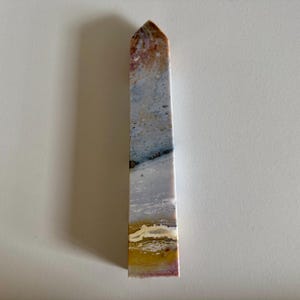 Op de afbeelding: Een rechthoekige stenen obelisk met een puntige top. De steen heeft een mix van kleuren, waaronder bruin, wit, grijs, geel en roze. De obelisk staat rechtop op een wit oppervlak.