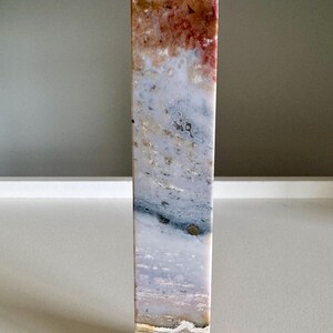 Op de afbeelding: Een hoge, rechthoekige stenen obelisk met een puntige top. De steen heeft een mix van kleuren, waaronder rood, bruin, wit en blauw, waardoor een gemarmerd effect ontstaat. De obelisk is een decoratief item.