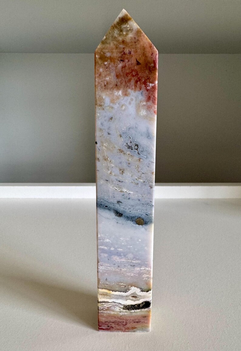 Op de afbeelding: Een hoge, rechthoekige stenen obelisk met een puntige top. De steen heeft een mix van kleuren, waaronder rood, bruin, wit en blauw, waardoor een gemarmerd effect ontstaat. De obelisk is een decoratief item.