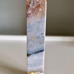 Op de afbeelding: Een hoge, rechthoekige stenen obelisk met een puntige top. De steen heeft een mix van kleuren, waaronder beige, roze, blauw en goud. De obelisk staat rechtop op een wit oppervlak.