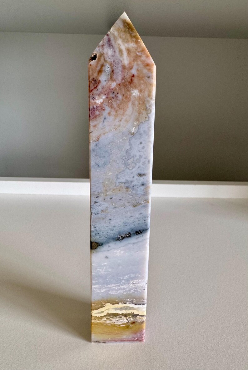 Op de afbeelding: Een hoge, rechthoekige stenen obelisk met een puntige top. De steen heeft een mix van kleuren, waaronder beige, roze, blauw en goud. De obelisk staat rechtop op een wit oppervlak.