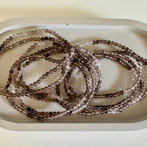 Op de afbeelding: Verschillende armbanden met kralen in bruine en heldere tinten zijn gerangschikt in een grijze, ovaalvormige schaal. De armbanden zijn gemaakt van kleine, ronde kralen, wat een delicate en elegante uitstraling creëert. De schaal biedt een neutrale achtergrond.