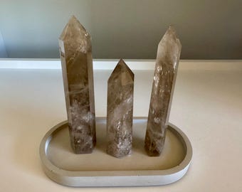 May include: Three smoky quartz crystal points of varying heights are displayed on a light gray, oval-shaped tray. The crystals have a translucent, brown hue with internal inclusions. The tray has a smooth, concrete-like texture.