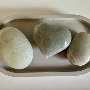 Op de afbeelding: Een grijze ovale betonnen tray bevat drie gepolijste stenen. Eén is hartvormig en de andere twee zijn ovaal. De stenen zijn in verschillende tinten grijs en wit. De tray heeft een lichtgrijze kleur.
