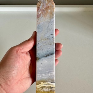 Op de afbeelding: Een rechthoekige kristallen obelisk met een puntige top. De steen heeft een mix van kleuren, waaronder beige, grijs en vleugjes geel en roze. De obelisk wordt in een hand gehouden, wat het gepolijste oppervlak en de natuurlijke patronen benadrukt.