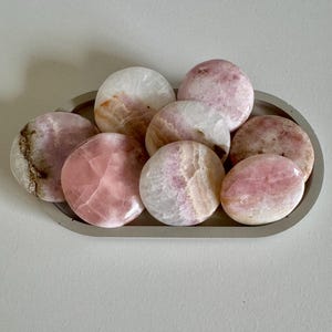 May include: A collection of polished, oval-shaped stones in shades of pink and white, arranged in a gray, oval tray. The stones have a smooth, glossy surface, and the color variations create a natural, organic aesthetic. The tray sits on a light gray surface.