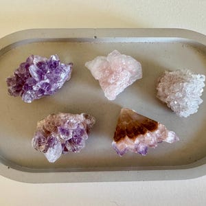 Op de afbeelding: Vijf ruwe kristallen in tinten paars, roze en wit zijn gerangschikt op een grijs ovaal dienblad. De kristallen variëren in grootte en vorm en tonen natuurlijke texturen en kleuren. De lade biedt een neutrale achtergrond.