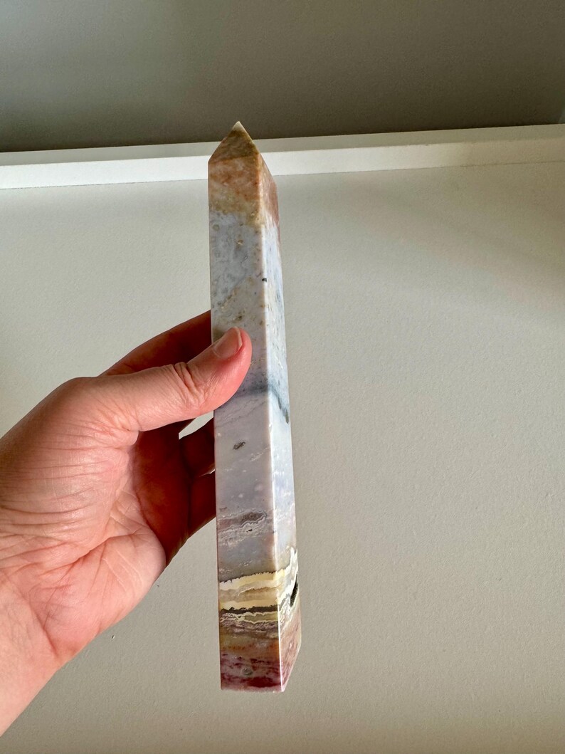 Op de afbeelding: Een hoge, rechthoekige kristallen obelisk met een puntige top. De steen heeft kleurstroken, waaronder wit, grijs, bruin en rood. De obelisk wordt in een hand gehouden.