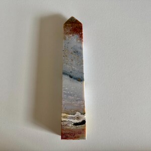 Op de afbeelding: Een hoge, rechthoekige stenen obelisk met een puntige top. De steen heeft een mix van kleuren, waaronder bruintinten, wit, grijs en zwart. De obelisk staat rechtop tegen een witte achtergrond en werpt een schaduw.